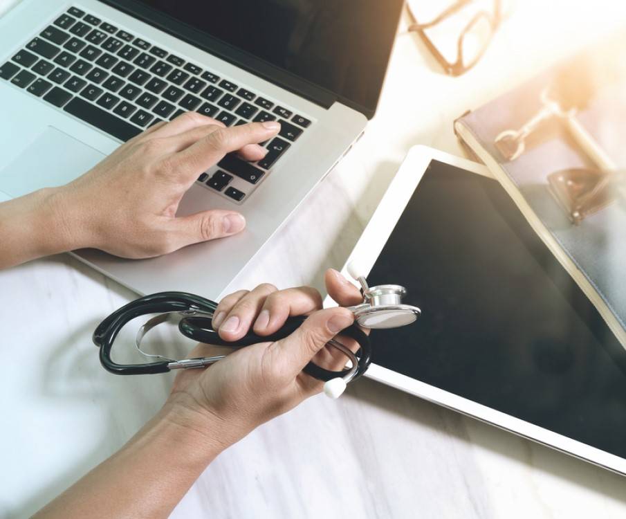 vrouw achter laptop met stethoscoop in haar hand en tablet op tafel